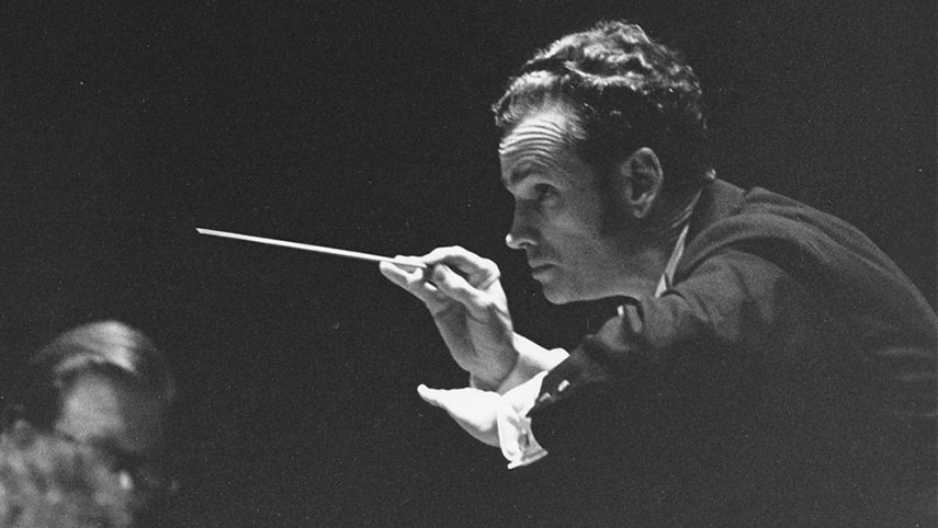 A close-up of Louis Lane conducting a concert at Severance Hall. He is bent forward slightly and gesturing to the players to play softly.