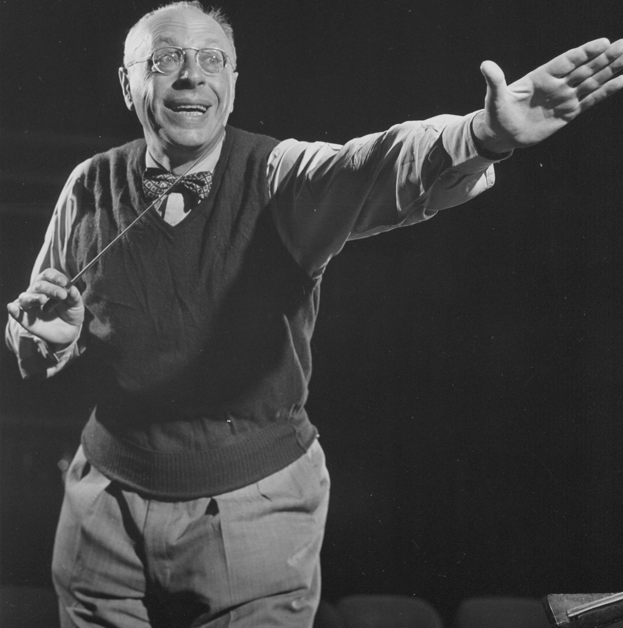 George Szell dressed in a sweater vest. Smiling, he excitedly points forward with his left hand. His right hand holds his baton. 