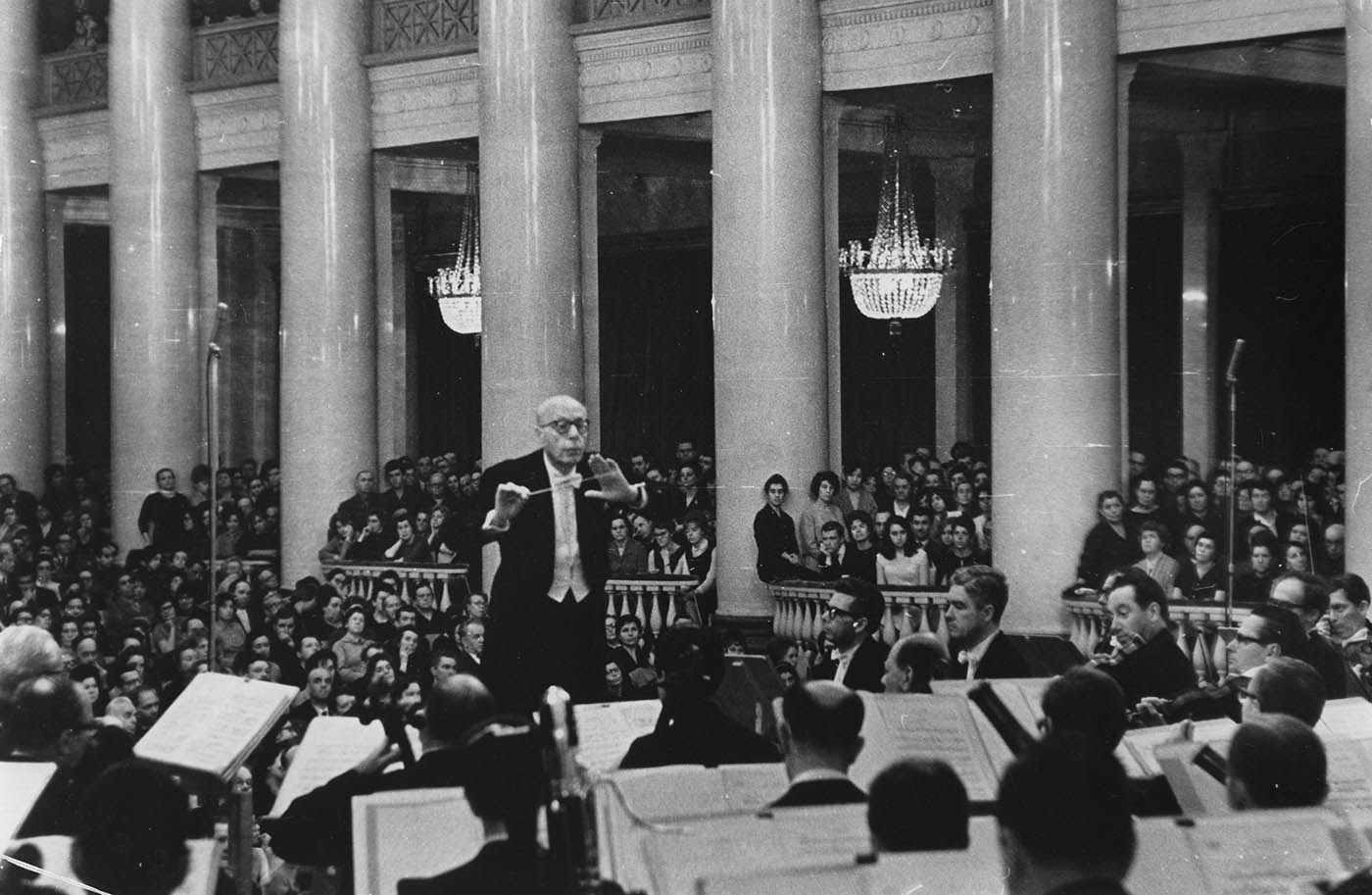 Szell is conducting the Orchestra. A large, attentive audience is seated behind them. Prominent chandeliers hang above the audience.  