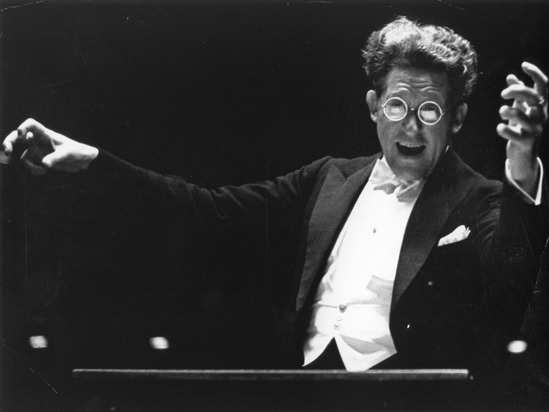 Artur Rodzinski conducting the Orchestra. He is facing the viewer, has a huge, excited smile on his face, and is wearing large glasses. 