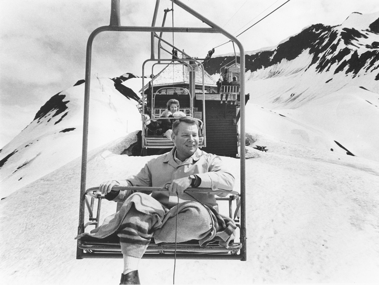A man on a ski lift, with mountains in the background.